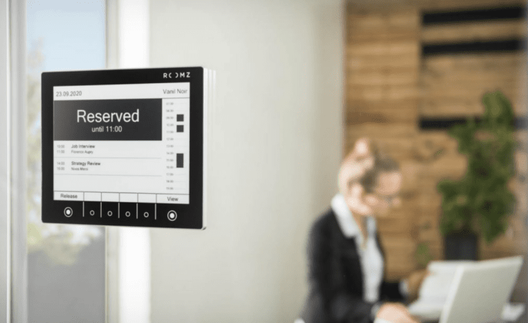 Ein ROOMZ-Display an einer Glaswand zeigt den Status „Reserved“ mit Uhrzeit und Rauminformationen an, um die Belegung von Besprechungsräumen transparent darzustellen.