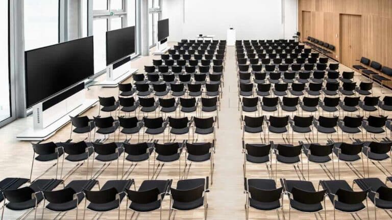 Ein großer, moderner Veranstaltungsraum mit Stuhlreihen, minimalistischer Architektur und einer Holzmedia S1 Medienwand mit großem Display – ideal für Konferenzen, Schulungen oder hybride Veranstaltungen mit professioneller Medientechnik.